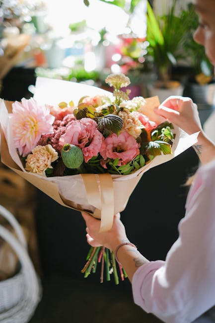 A person holding a large bouquet of fresh flowers, including pink peonies, light yellow carnations, and small white and purple blooms, wrapped in white and beige paper with a cream-colored ribbon. The