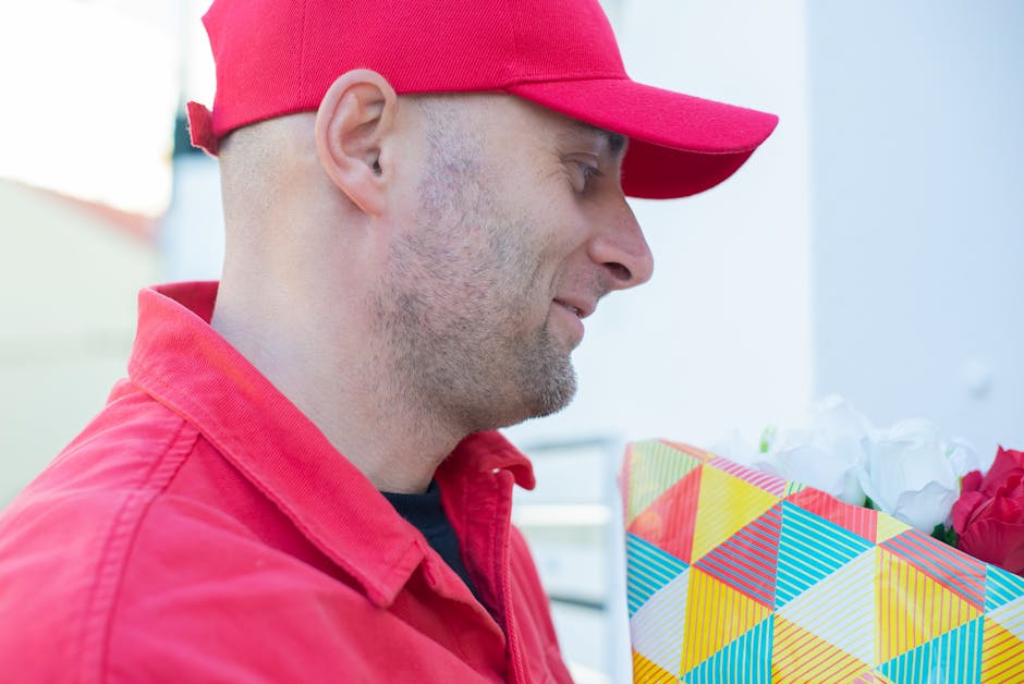 A man dressed in a red cap and red jacket holds a colorful, geometric-patterned gift bag filled with white and pink flowers, featuring roses and other blooms, set against a bright, softly blurred outd