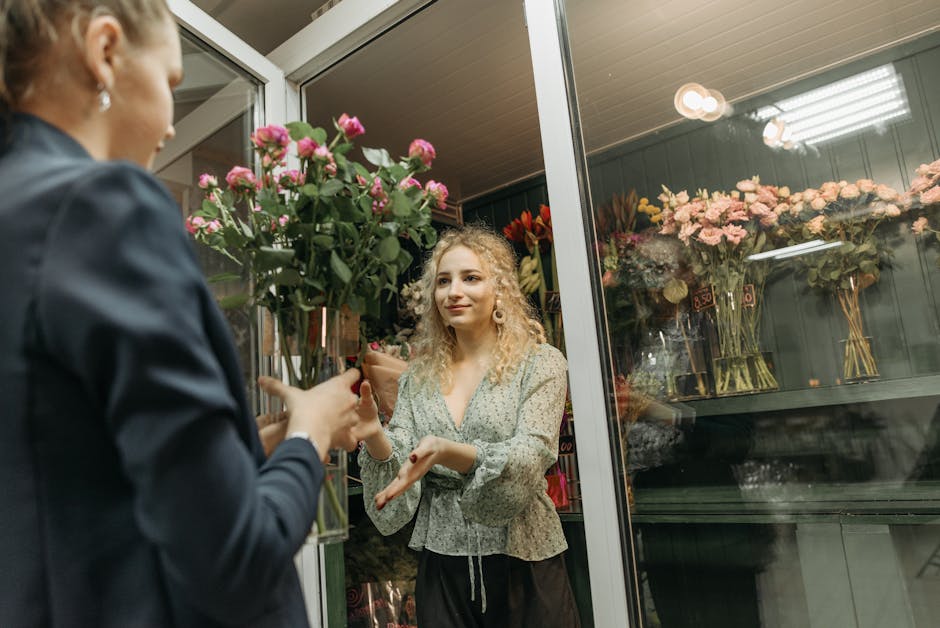A florist with blonde, curly hair and a floral-patterned blouse is handing over a bouquet of pink roses wrapped in brown paper to a customer. The arrangement features fresh, vibrant pink roses with gr