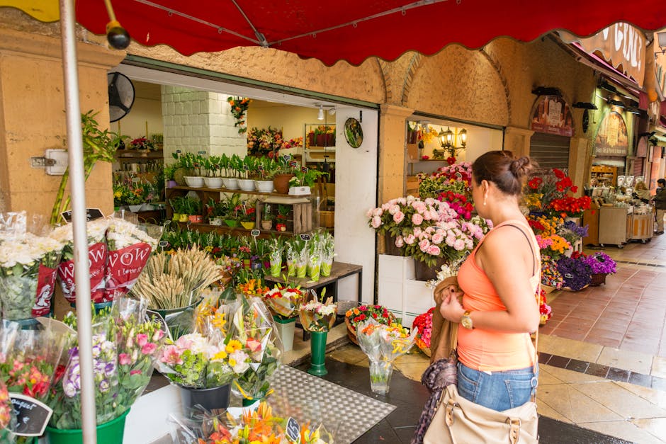 A woman with brown hair tied in a bun, wearing an orange sleeveless top and blue jeans, stands in front of a flower shop under a red canopy. She is holding a small bouquet of pink and red roses wrappe