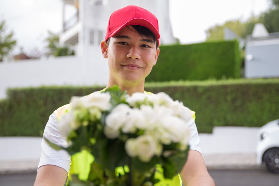 A delivery person wearing a red cap and high-visibility vest is smiling warmly while holding a bouquet of fresh white flowers, including roses and lilies, wrapped in clear cellophane with green foliag