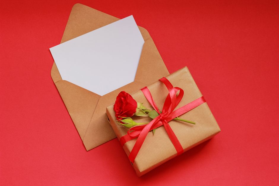 A neatly wrapped gift box with brown paper and a red satin ribbon tied in a bow, adorned with a single red rose and green foliage, is placed next to an open brown kraft envelope with a blank white car