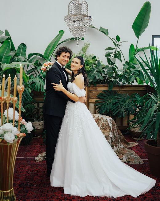 A newlywed couple stands together indoors, surrounded by lush green tropical plants and a vintage chandelier hanging above. The groom is dressed in a classic black tuxedo, while the bride wears a whit