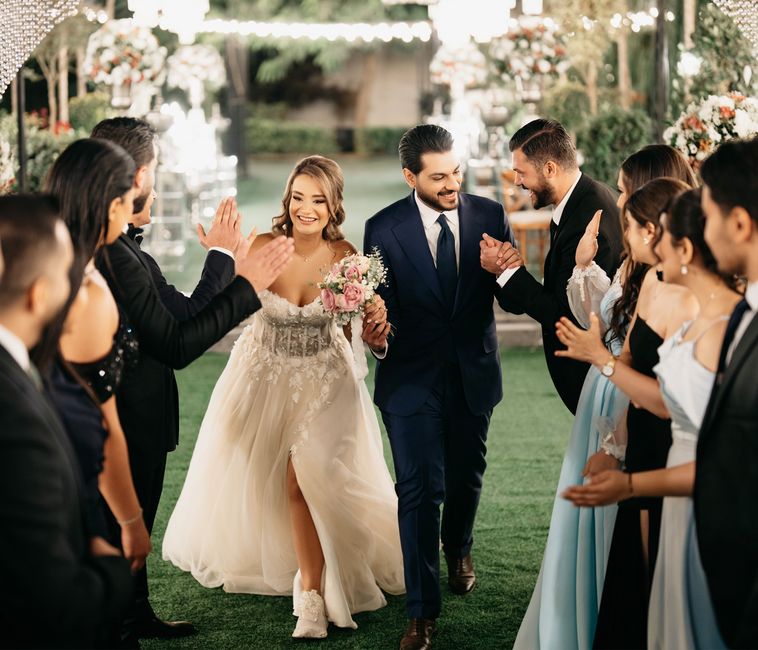 A bride in a lace and tulle wedding gown, holding a bouquet of pink roses, peonies, and white filler flowers, is walking hand-in-hand with a groom dressed in a navy blue suit and white shirt. They are