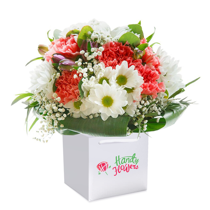 A masterfully composed bouquet, captured in luminous detail, stands tall within a square white gift bag-an embodiment of Mortlake's subtle sophistication and neighbourhood warmth. The composition brings together lush coral and soft peach carnations, their frilled petals suggesting both liveliness and sureness of hand. These are paired with pristine white daisies and daisy-like chrysanthemums, each glowing with a lime-green heart that adds a refreshing burst of contrast. Scattered throughout are diminutive clusters of gypsophila, their snowflake-like blossoms softening each transition between colour and petal. At the left, the appearance of a few closed, purple-tipped buds injects a sense of quiet suspense, echoing the hidden gem gardens tucked along Church Avenue. The bouquet is ringed by broad, gleaming green leaves, which not only support the stems but also create a gently curving cradle that guides the eye around the arrangement's rounded shape. The crisp white gift bag, complete with ribbon handles for easy carrying or delivery along Upper Richmond Road, presents the bouquet as a ready-to-give treasure. Soft natural lighting showcases every hue and contour, hinting at the bouquet's sweet fragrance and the meticulous care taken by Mortlake Florist. Whether chosen for a special milestone, an impromptu act of kindness, or a cherished holiday, this design radiates abundant freshness and the inviting elegance of its riverside locale.