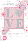 This anniversary card presents a gentle celebration of enduring romance, wrapped in soft pastel tones and delicate shine. Center stage sits the word LOVE, formed from raised lettering dusted with fine, blush-pink glitter, while the letter O is subtly hinted as a heart shape, edged in shimmering silver that glows against the powder-pink background. The surface looks almost velvety to the touch, with the glitter catching the light like tiny reflections on the Thames near Mortlake. Surrounding this focal point, clusters of illustrated blossoms in blush, mauve, and pale ivory unfurl with a watercolour softness, their outlines captured in fine, flowing lines that loop and curl like trailing vines in a well-tended garden. Small flourishes of petals, swirls, and tiny, almost-hidden butterflies drift around the flowers, suggesting the same quiet grace you might find in a thoughtfully arranged bouquet from Mortlake Florist on Sheen Lane. The background blush gently deepens and lightens in places, conveying depth without losing its calm, romantic feel. A softly scripted message-phrases like On Our Anniversary and I love you today and always-rests beneath or above the floral motif in an understated grey script, easy to read yet unobtrusive. The overall mood is tender and intimate, like a private promise shared at home after an evening walk past Mortlake Green. This card looks as if it was designed to be paired with fresh roses and seasonal flowers, ready for local anniversary delivery across Mortlake and the surrounding riverside streets.